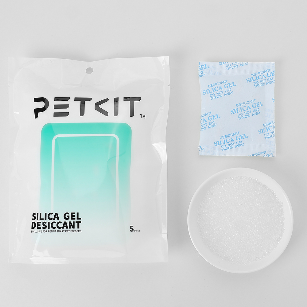 Baltas PETKIT pakuotė su juodu logotipu viršuje ir žalsvai mėlynu stačiakampiu centre. Šalia pakuotės – trys nedideli permatomi maišeliai su silikagelio granulėmis ir baltas dubenėlis, pripildytas baltų silikagelio granulių.